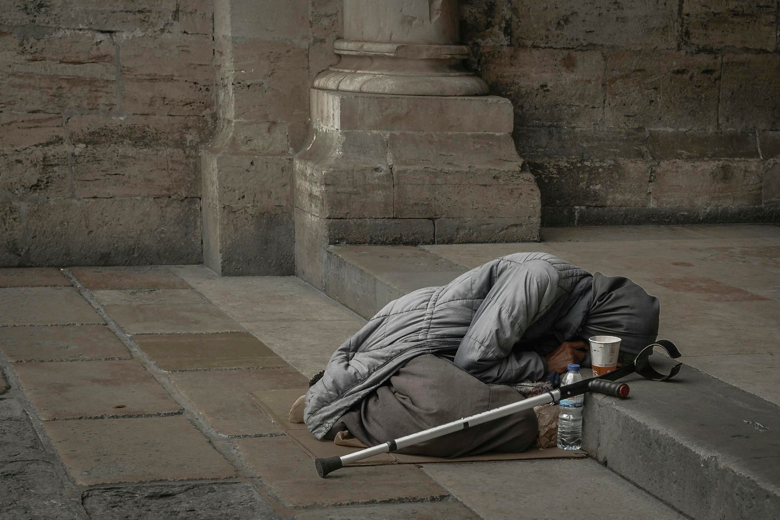 A homeless woman in a gray jacket with a hijab rests against a stone wall, highlighting urban poverty.